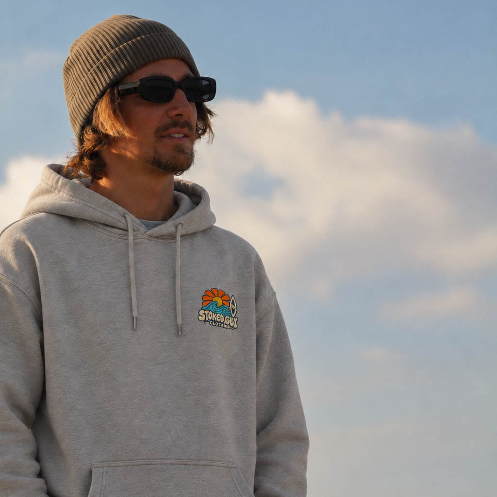 A man wearing a light gray heather hoodie with a colorful 'Palm Springs' graphic on the left chest, a brown knit beanie, and sunglasses, standing outdoors against a blue sky with clouds.