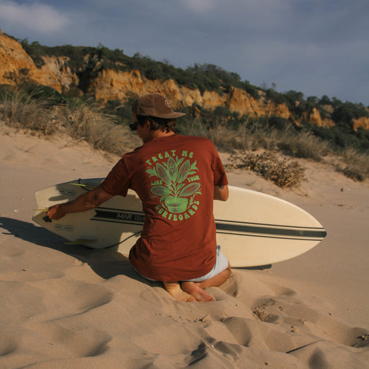 Person sitting on a sandy beach with a surfboard, wearing a red t-shirt with a green graphic.