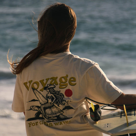 Person holding a surfboard with 'Voyage' t-shirt on a beach