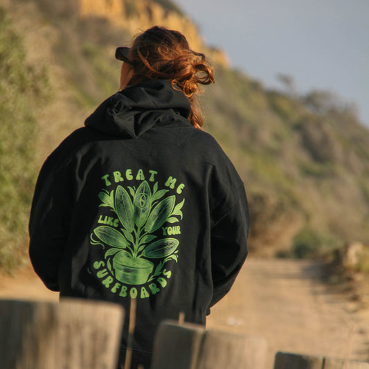 Person wearing a black hoodie with a green graphic and text, standing on a wooden bridge with a scenic background.