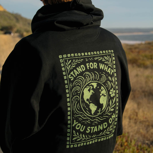 Person wearing a black hoodie with a green graphic and text on the back, standing in a natural landscape.