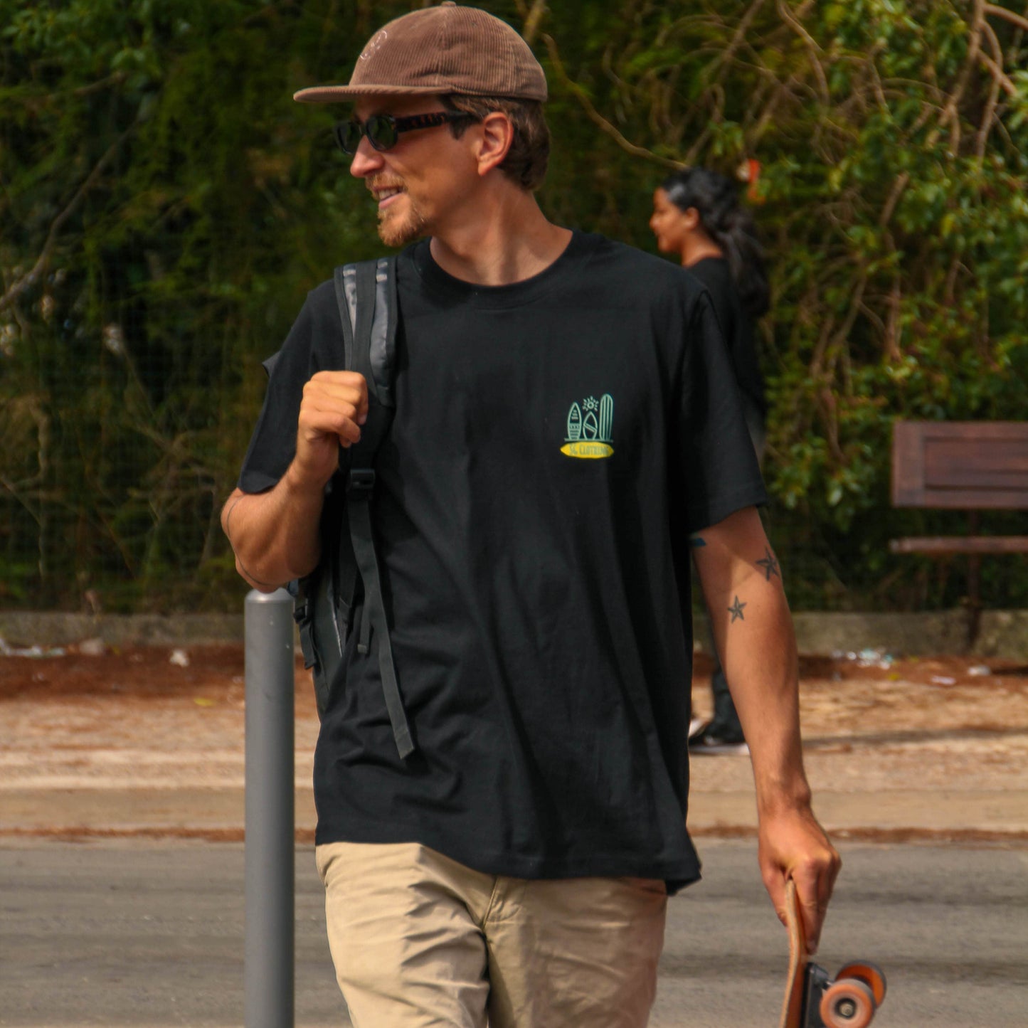 Man walking with a skateboard, wearing a black t-shirt and beige shorts, in an outdoor setting.