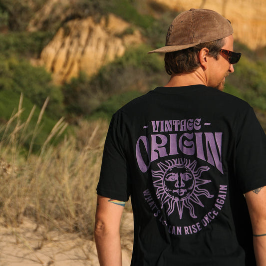 Person wearing a black t-shirt with purple text and graphics, standing on a beach with cliffs in the background.