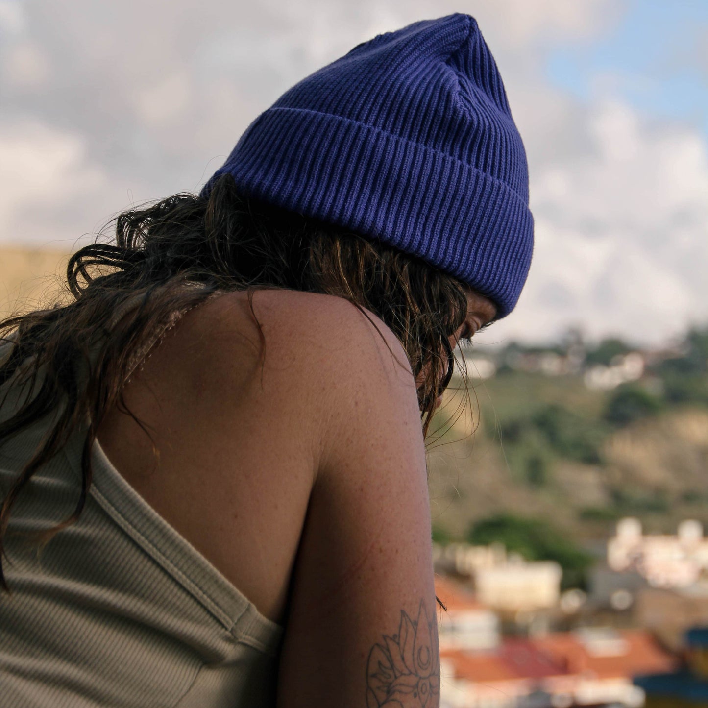 Person wearing a blue beanie looking over a scenic landscape with buildings and greenery.
