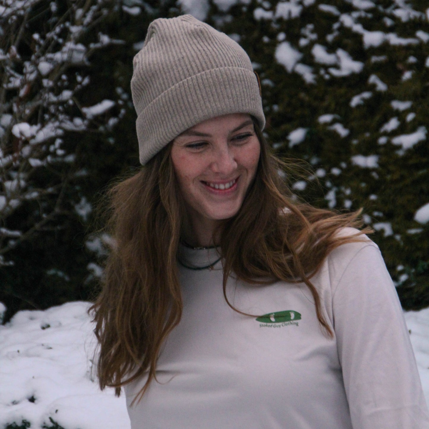 Person wearing a beige knit hat and light-colored jacket standing in a snowy landscape with trees.