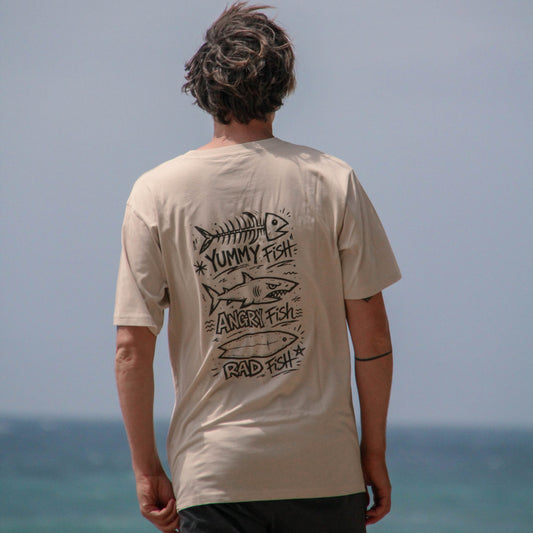 Person wearing a beige t-shirt with graphic designs and text, standing on a beach.