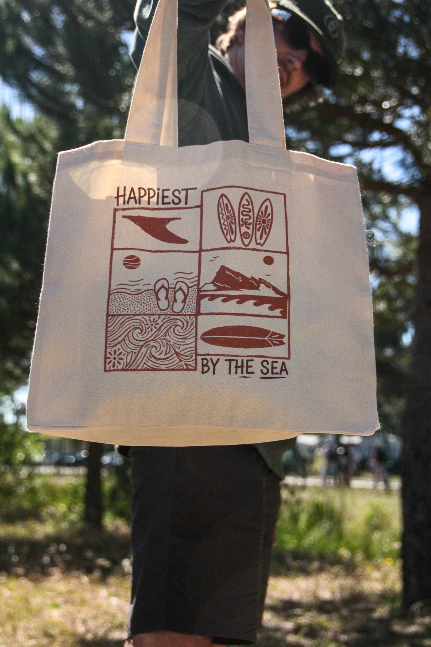 Person holding a tote bag with 'Happiest by the Sea' design outdoors.