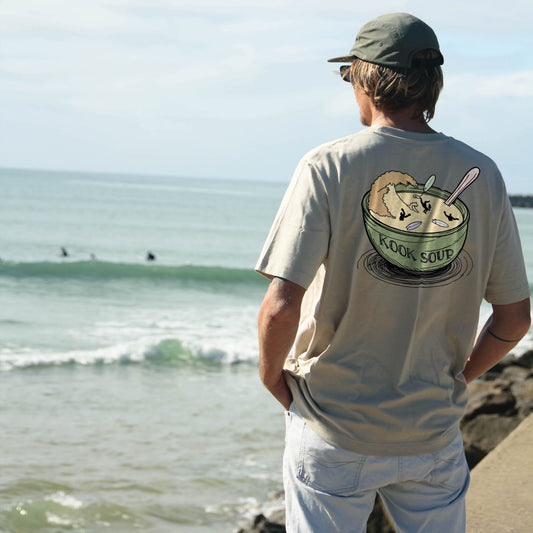 A beige colored organic cotton t-shirt with a graphic print of a bowl of soup with the text 'KOOK SOUP' and a spoon in it, and a smaller graphic of a person's face on the upper left chest area.