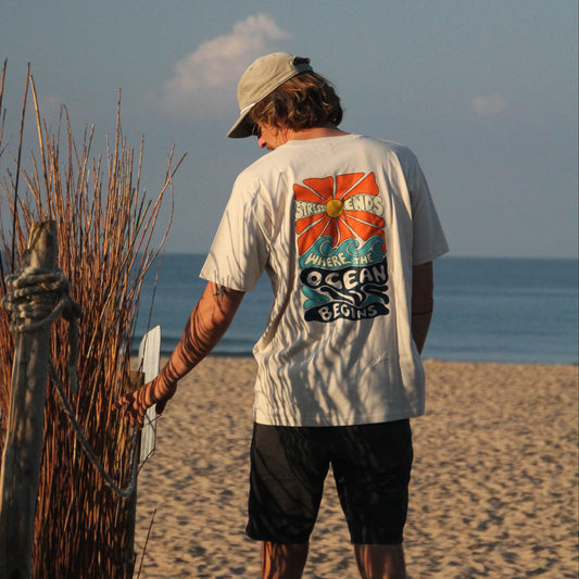 A man standing on a sandy beach facing the ocean, wearing a white graphic t-shirt with a colorful 'Ocean' print on the back, black shorts, and a beige cap.