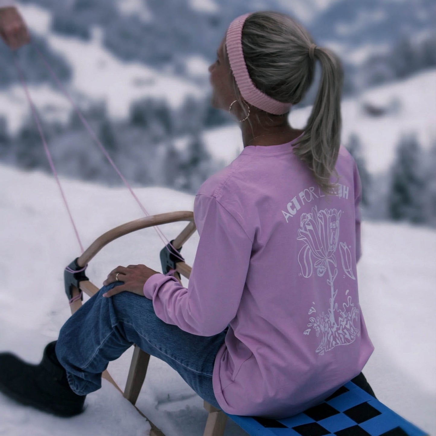 Person in pink shirt and blue jeans sitting on a sled in a snowy mountain landscape