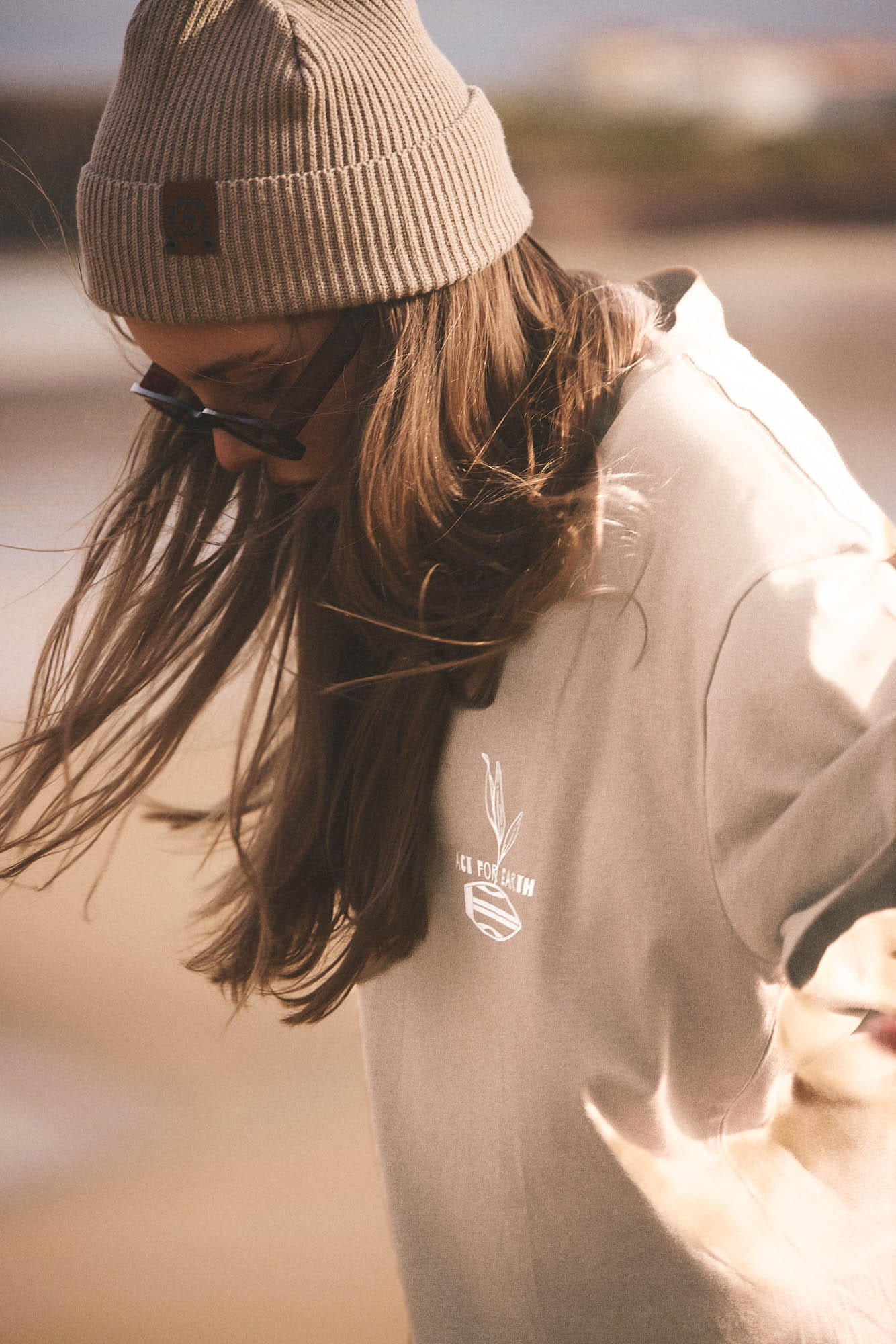 A person wearing a beige ribbed beanie, sunglasses, and a light beige sweatshirt with a small white chest graphic, photographed outdoors in warm, backlit sunlight.