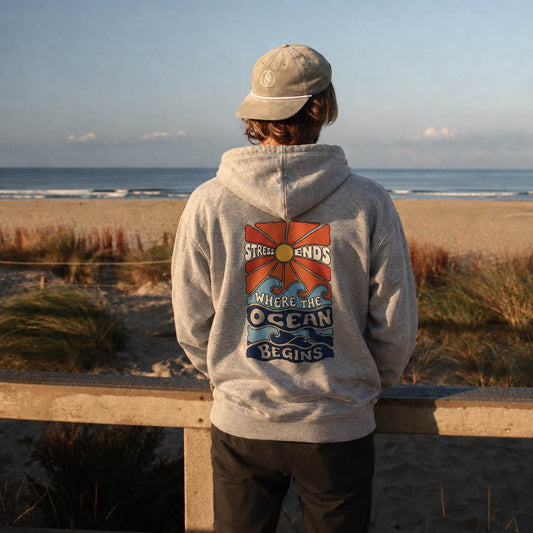 A person standing on a beach dune overlooking the ocean, wearing a light gray hoodie with a large back graphic that reads 'ST. HELENA'S OCEAN BEGINS' and a beige cap.