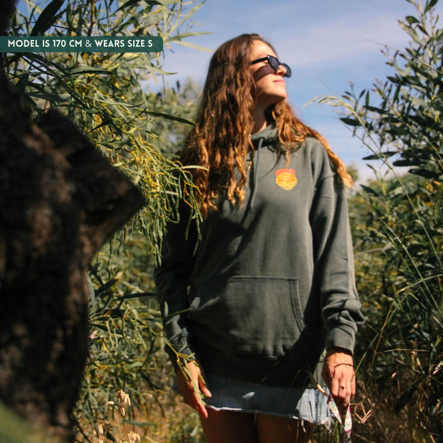 Woman wearing a dark green hoodie with a logo, standing in an outdoor setting with trees.