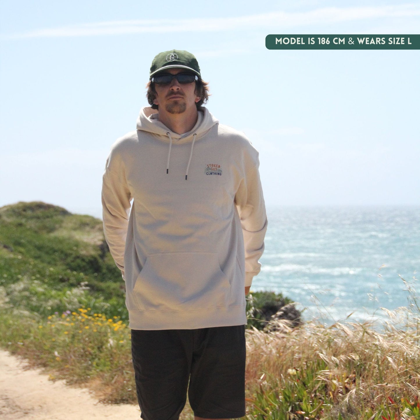 Man wearing a white hoodie and green cap standing on a coastal path with ocean view