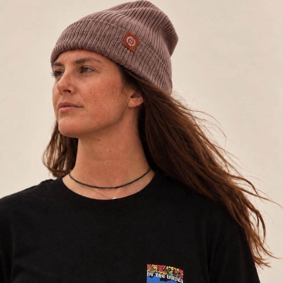 A woman wearing a ribbed mauve beanie with a small circular logo patch and a black crewneck sweatshirt with a colorful chest graphic, photographed against a plain light background.