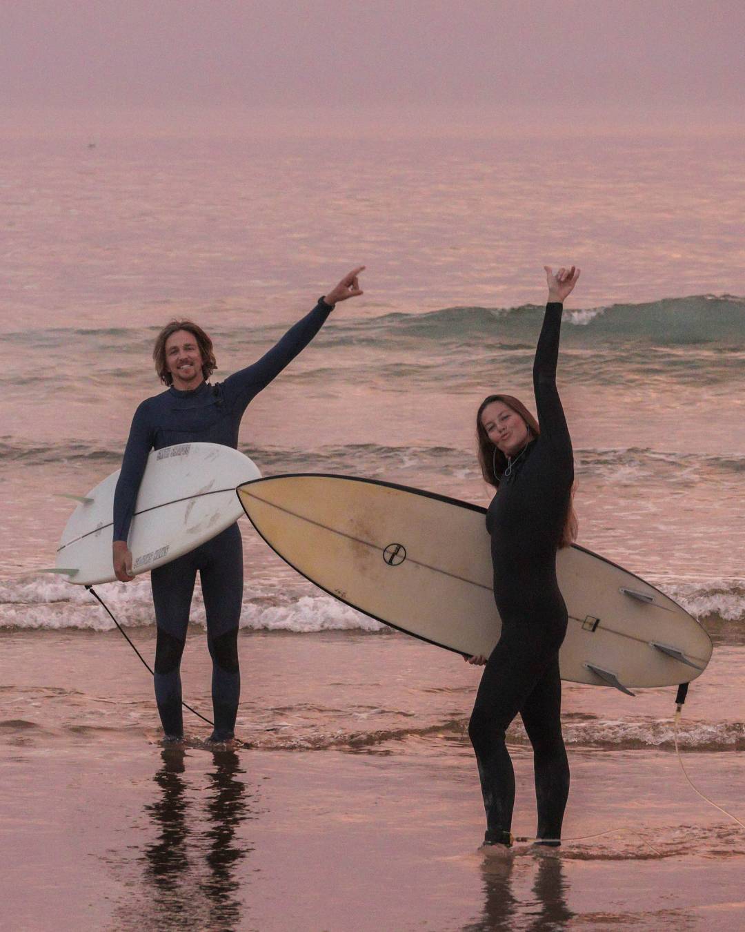 Two surfers in full-body wetsuits standing in shallow ocean water at sunset, each holding a surfboard and raising one arm. The sky and water have a pinkish hue, and no brand logos are visible.
