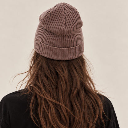 A person viewed from behind wearing a ribbed knit beanie hat in a muted mauve-brown shade, with long wavy brown hair and a black top against a plain light beige background.