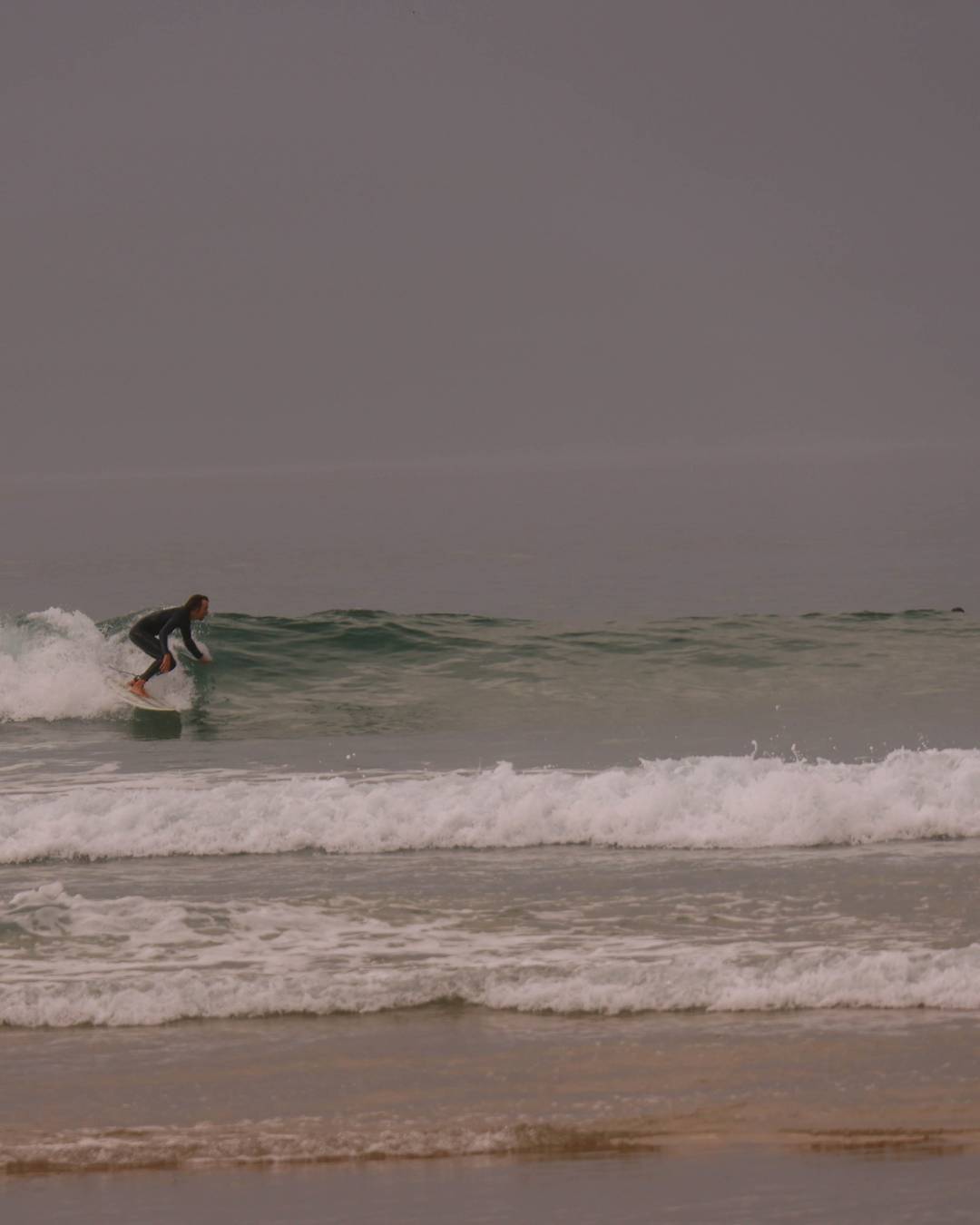 A surfer in a dark wetsuit rides a small wave on a surfboard at a beach, with white foam lines and a muted gray sky above.