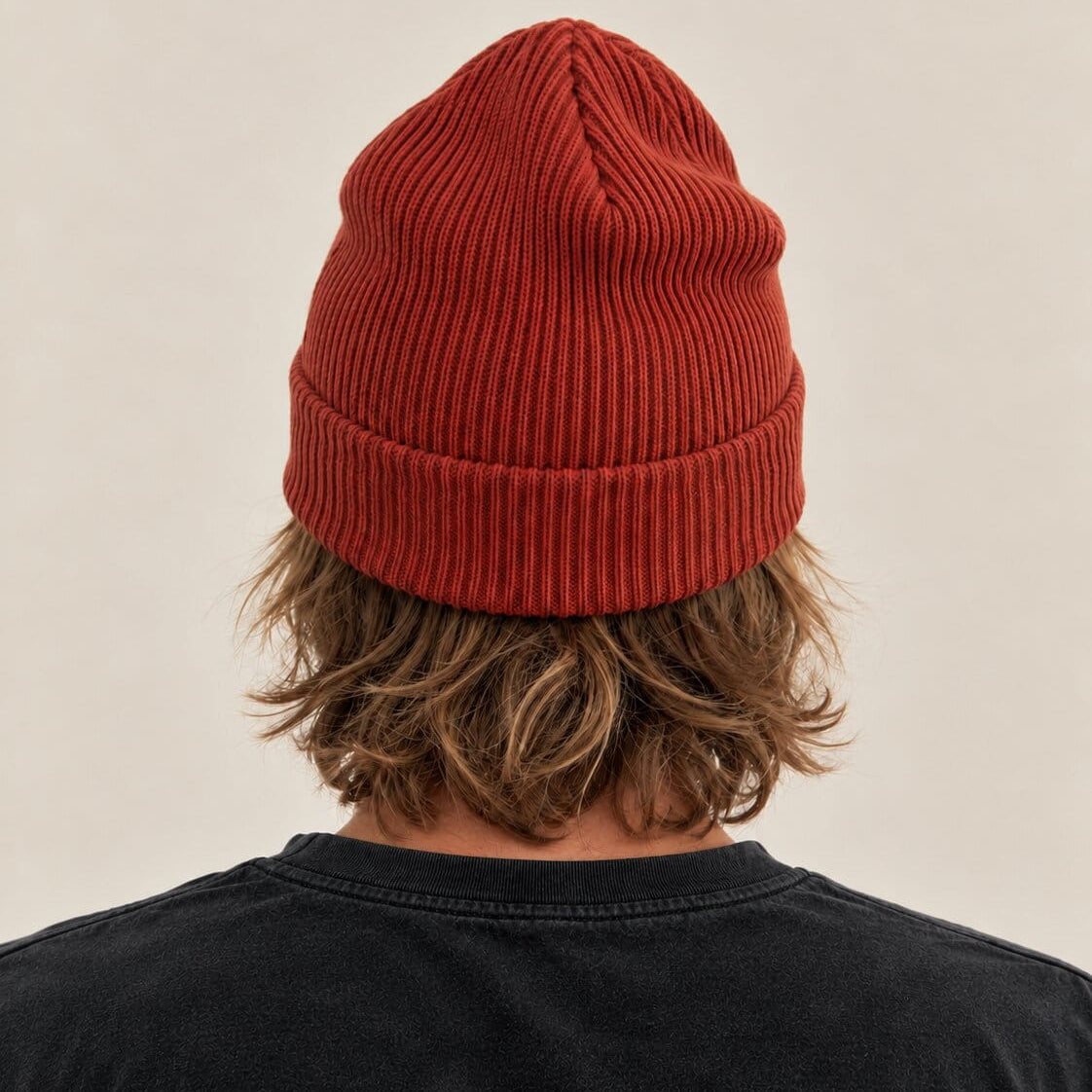 A person viewed from behind wearing a rust-red ribbed knit beanie and a black t-shirt against a plain beige background.