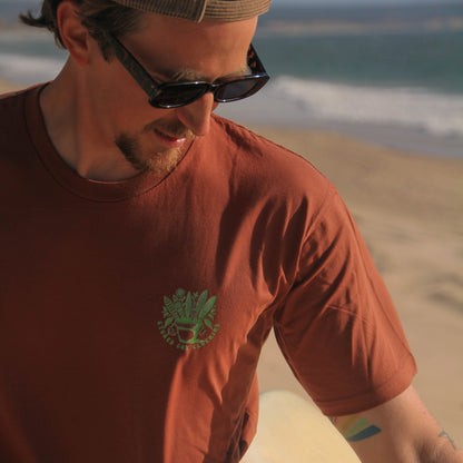 Man in a red t-shirt with a logo, sunglasses, and a cap holding a surfboard on a beach.
