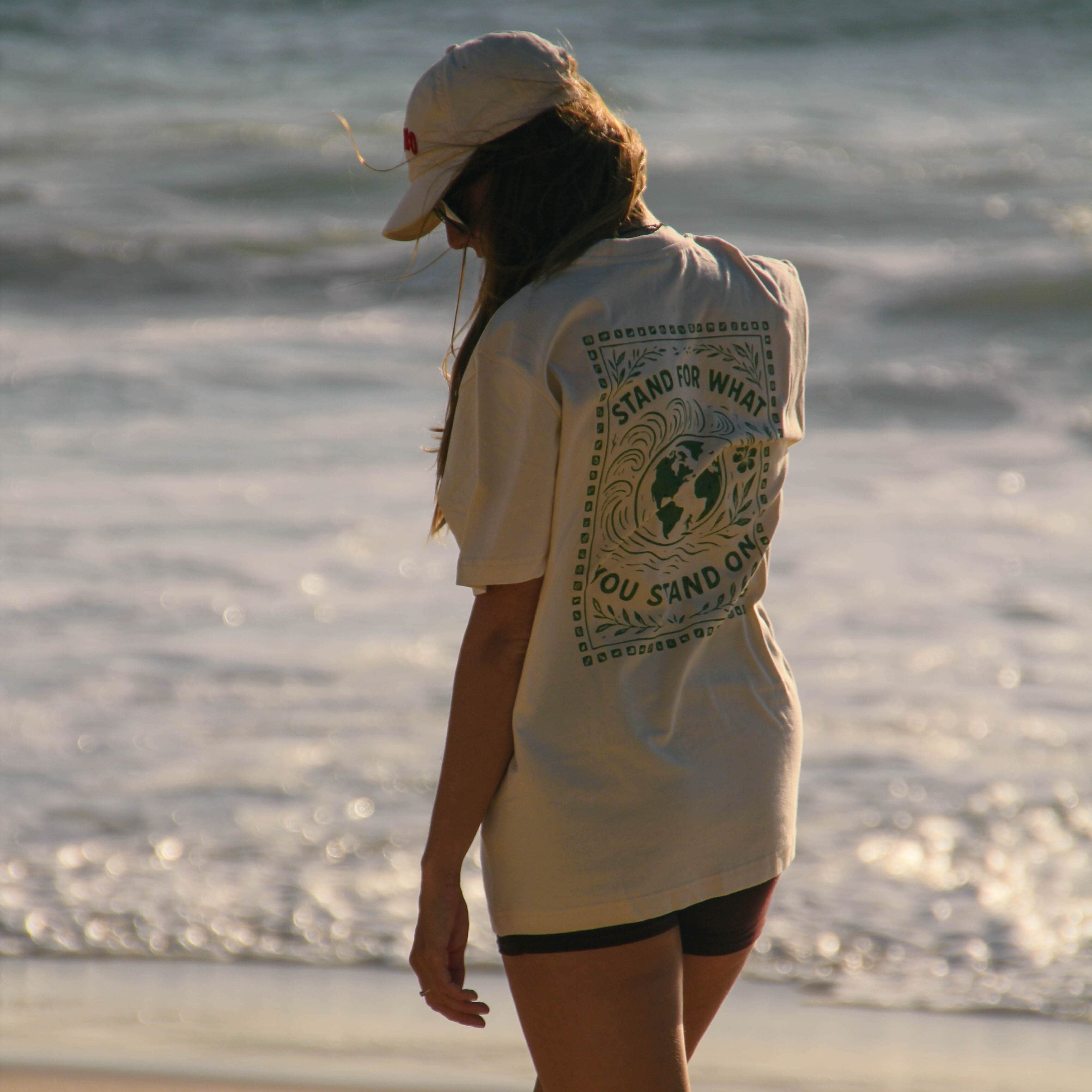 Person walking on a beach wearing a white t-shirt with graphics and text.