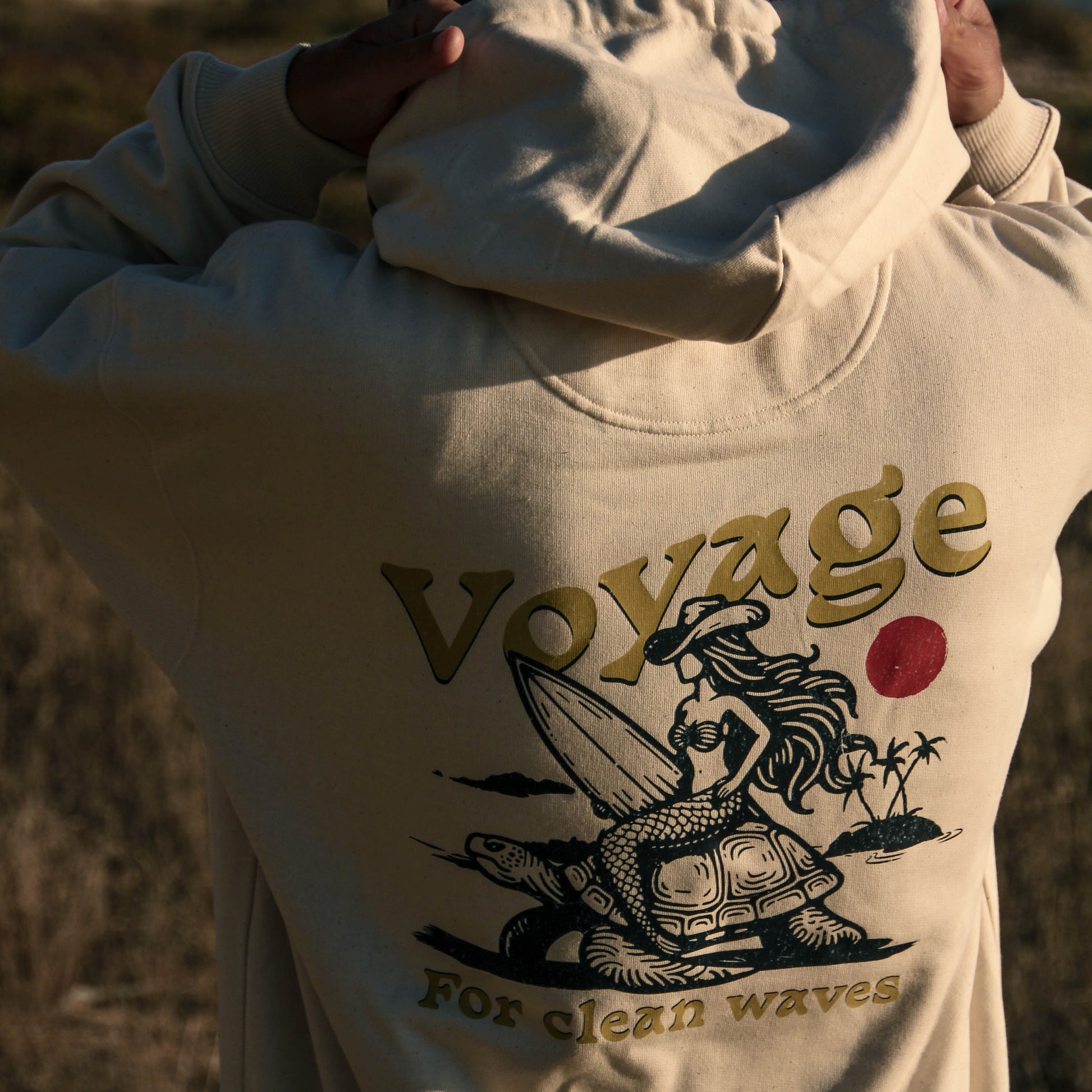 Person wearing a hoodie with 'Voyage for clean waves' design by the beach.