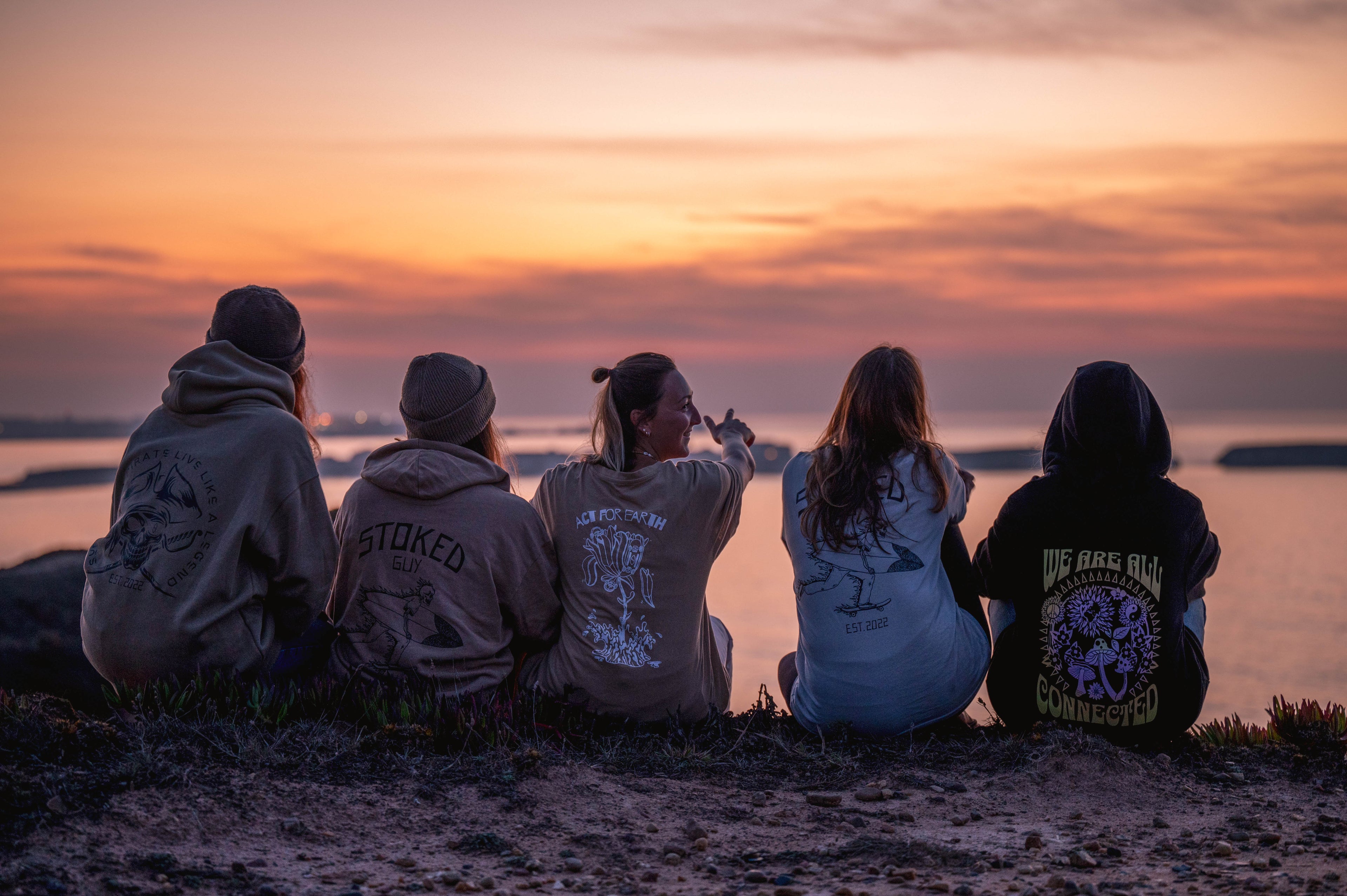 Five people sitting on a beach at sunset, facing the ocean.