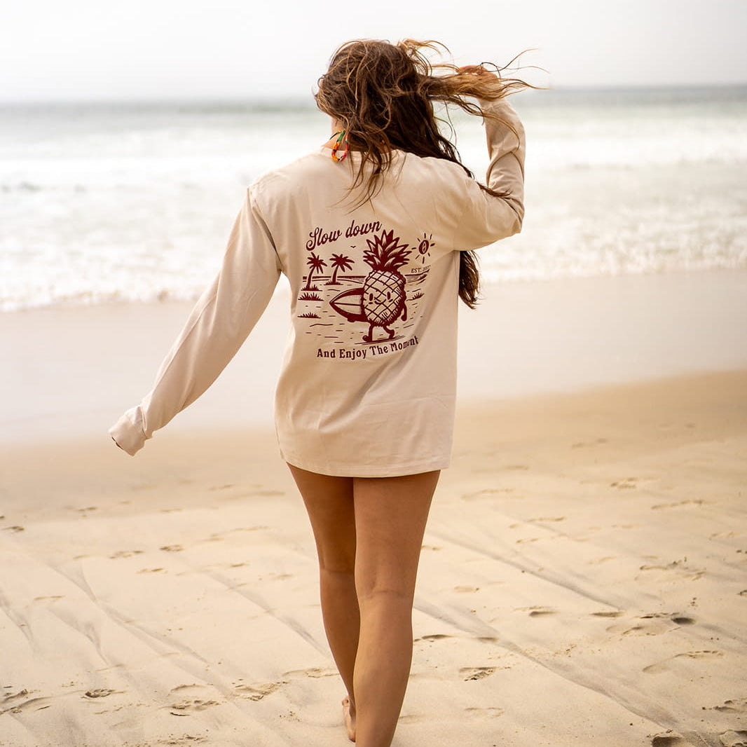 Woman walking on a beach wearing a beige long-sleeve shirt with a pineapple graphic.