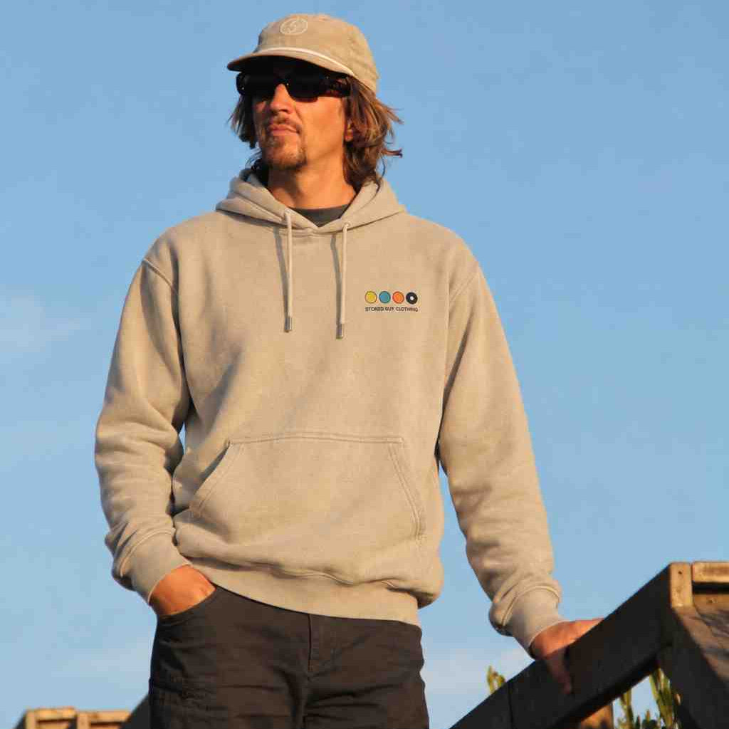 A man wearing a light beige hoodie with a small colorful 'sooo' logo on the left chest, a matching beige cap, and dark pants, standing outdoors against a clear blue sky.