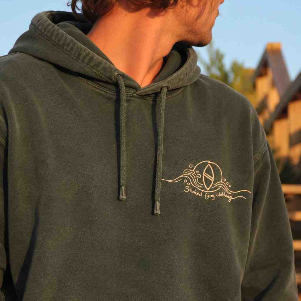 A person wearing a dark olive green hooded sweatshirt with a beige graphic print on the left chest, photographed outdoors in warm sunlight.