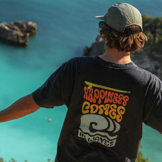 Person wearing a t-shirt with 'Happiness Comes in Waves' design overlooking a turquoise ocean.