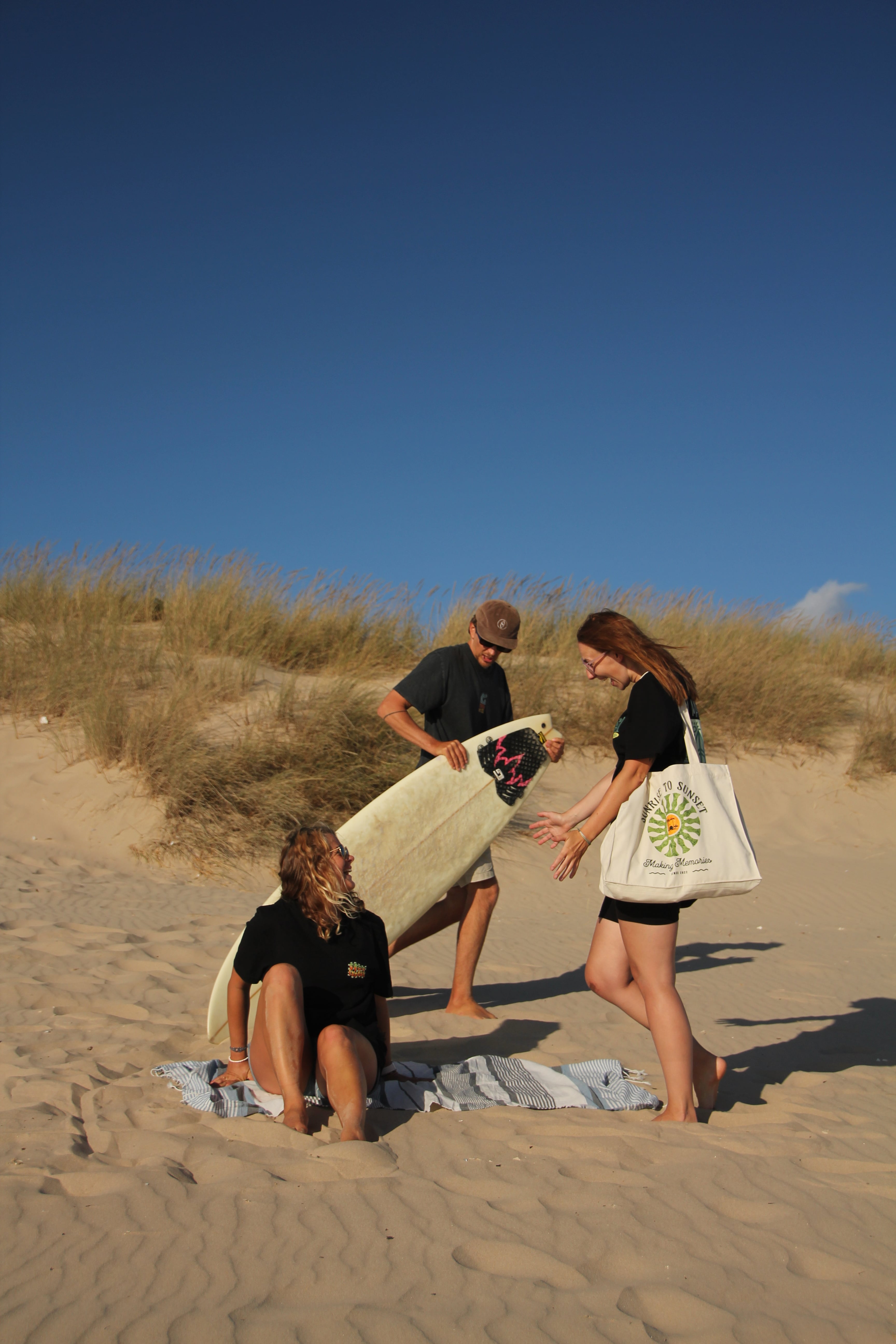 Three people on a sandy beach with one person holding a surfboard and another carrying a white tote bag.