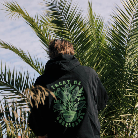 Person wearing a black hoodie with a green leaf design, standing among palm trees against a blue sky.