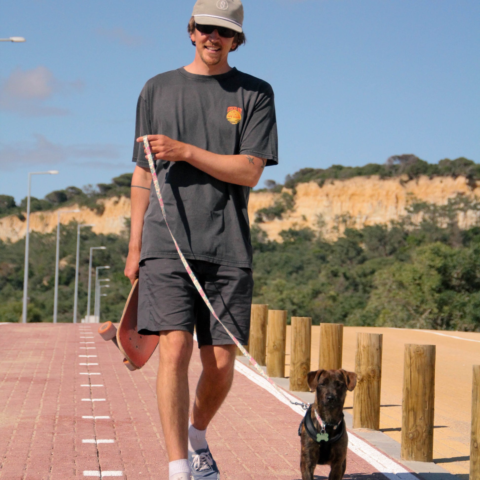Man walking a dog on a leash with a skateboard under his arm, set against a scenic background.