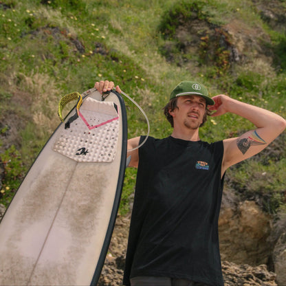 Man holding a surfboard with a green cap and black shirt in a natural setting