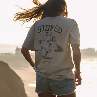 A woman stands on a beach cliff at sunset wearing a cream graphic t-shirt with 'STOKED GUY' and a surfer illustration on the back, paired with denim shorts.