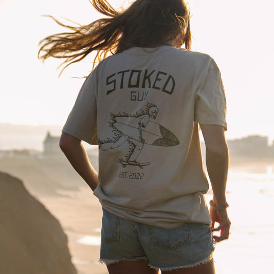 A woman stands on a beach cliff at sunset wearing a cream graphic t-shirt with 'STOKED GUY' and a surfer illustration on the back, paired with denim shorts.