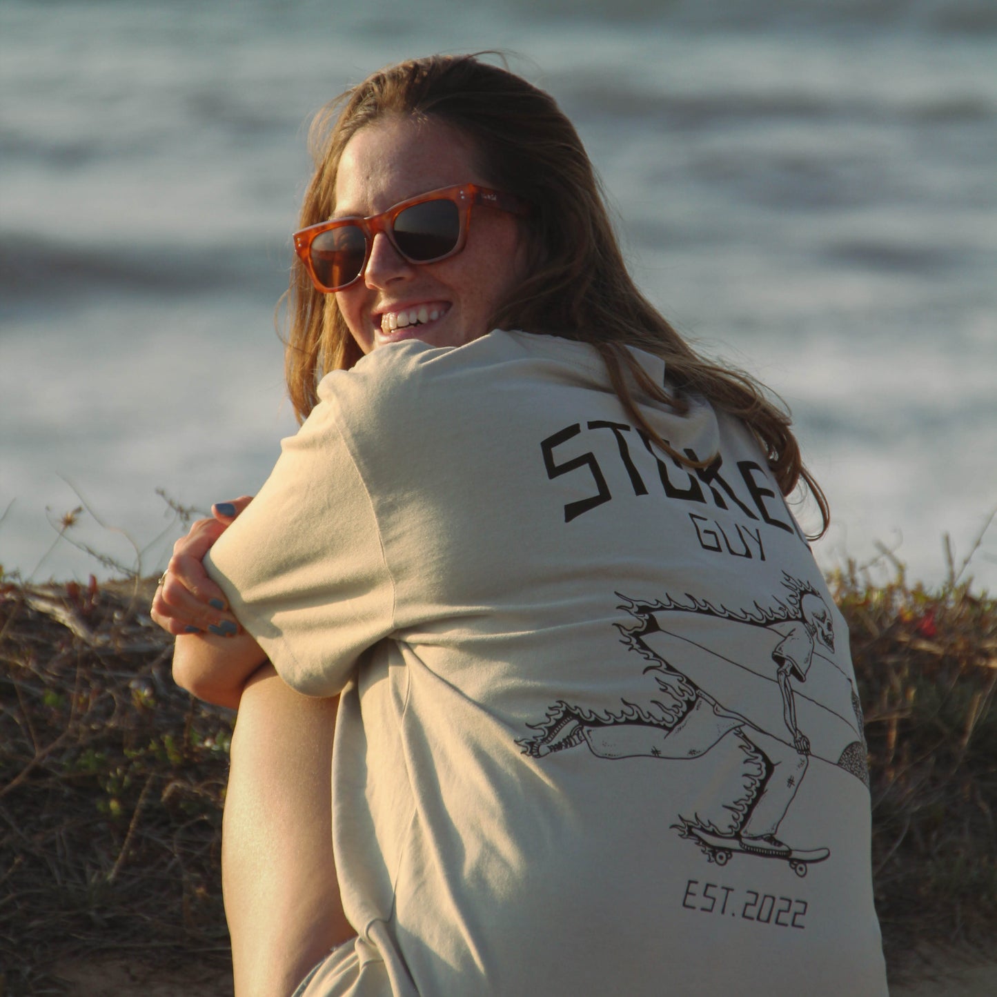 A woman sitting on a beach wearing a cream-colored graphic t-shirt with 'STICKER GUY EST. 2022' printed on the back, along with orange-tinted sunglasses.
