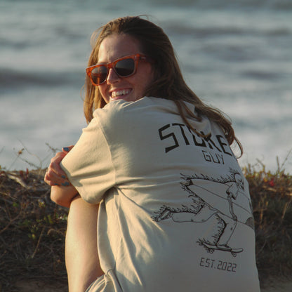 A woman sitting on a beach wearing a cream-colored graphic t-shirt with 'STICKER GUY EST. 2022' printed on the back, along with orange-tinted sunglasses.