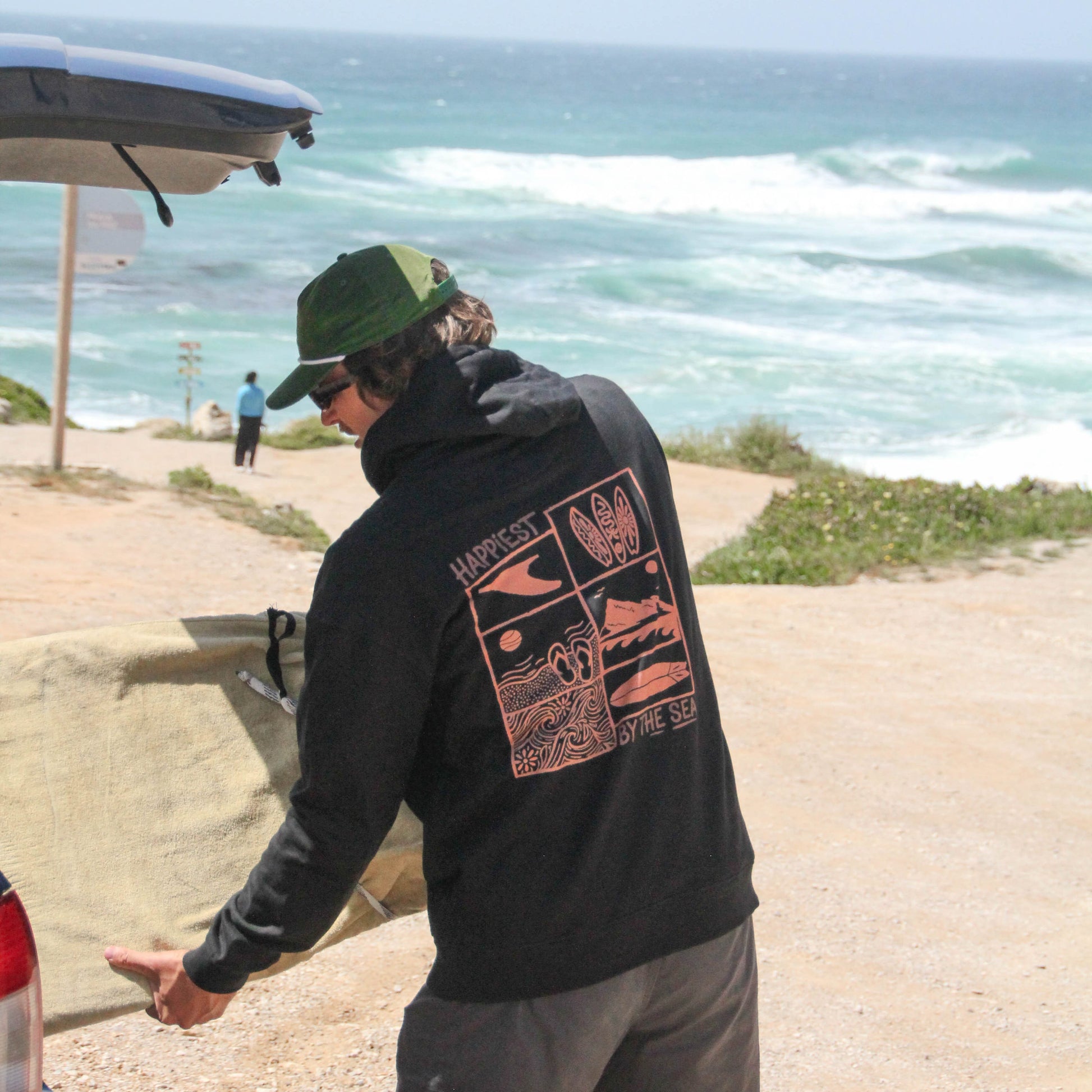 Person wearing a black hoodie with a graphic design, standing by a coastal road with ocean view.