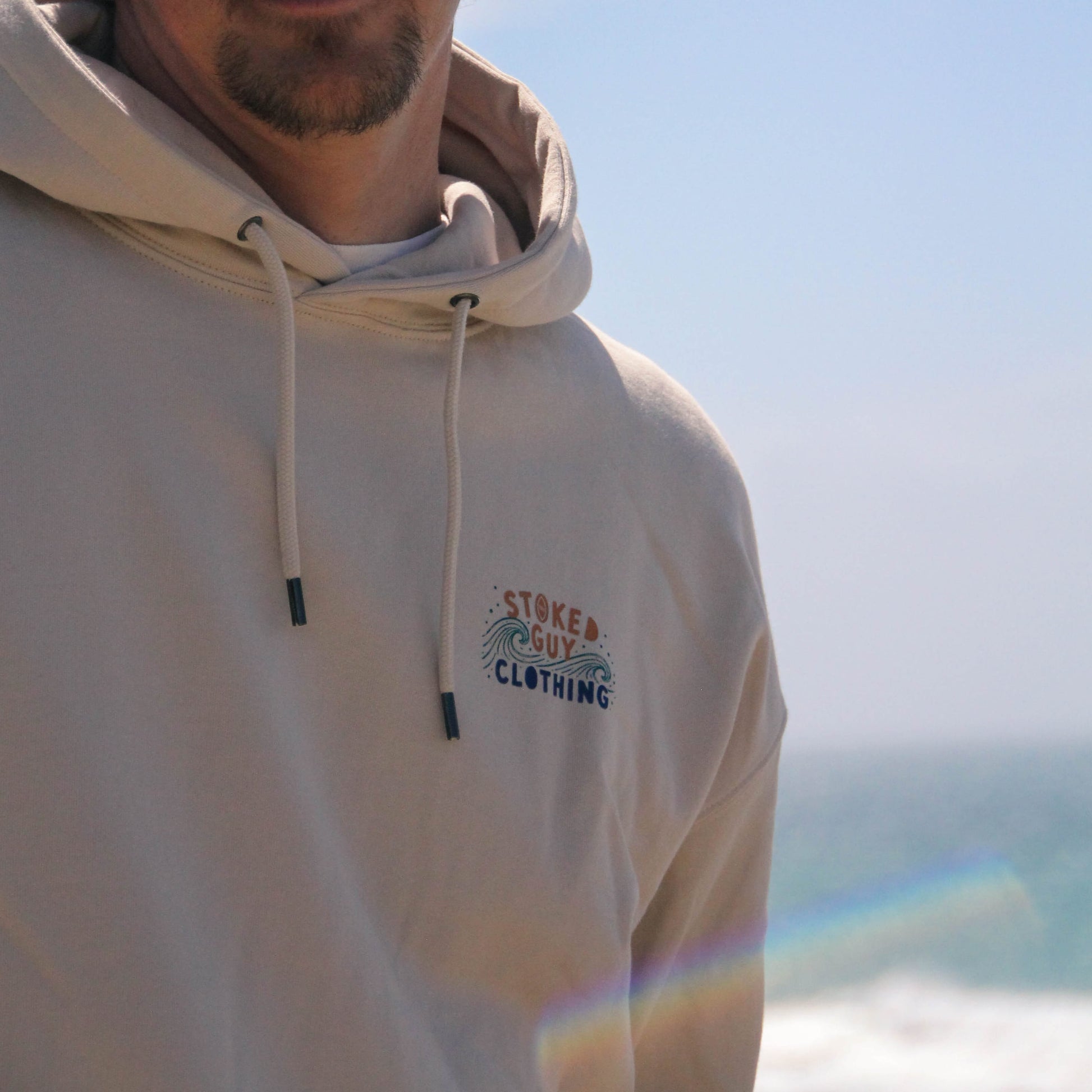 Person wearing a beige hoodie with 'Stoked Guy Clothing' logo against a blurred natural background