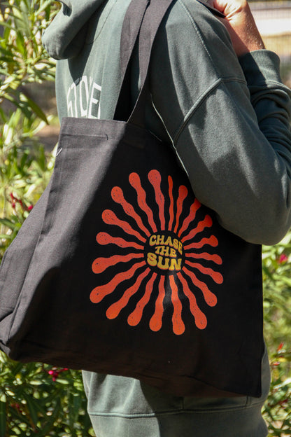 Person holding a black tote bag with a red and yellow design and text 'Chase the Sun'.
