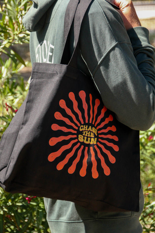 Person holding a black tote bag with a red and yellow design and text 'Chase the Sun'.