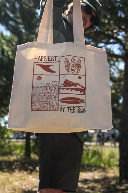 Person holding a tote bag with 'Happiest by the Sea' design outdoors.