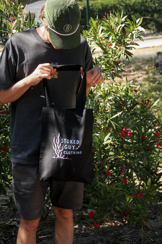Person holding a black tote bag with 'Stoked Guy Clothing' logo outdoors.