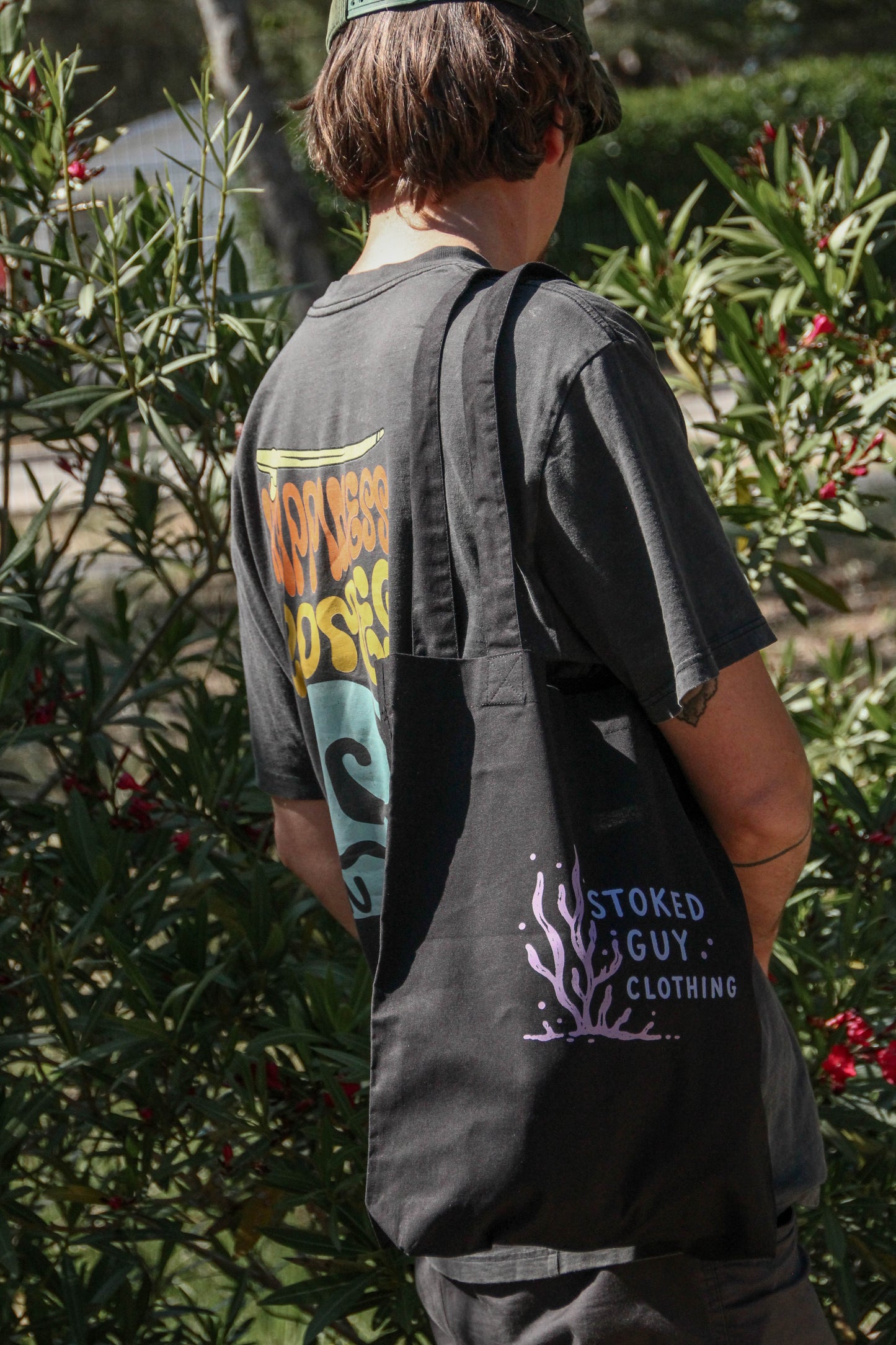 Person wearing a black tote bag with branding, standing among greenery.