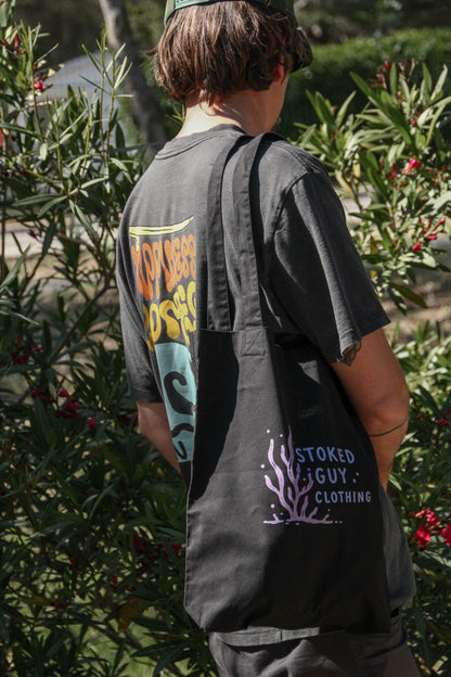 Person wearing a black tote bag with branding, standing among greenery.