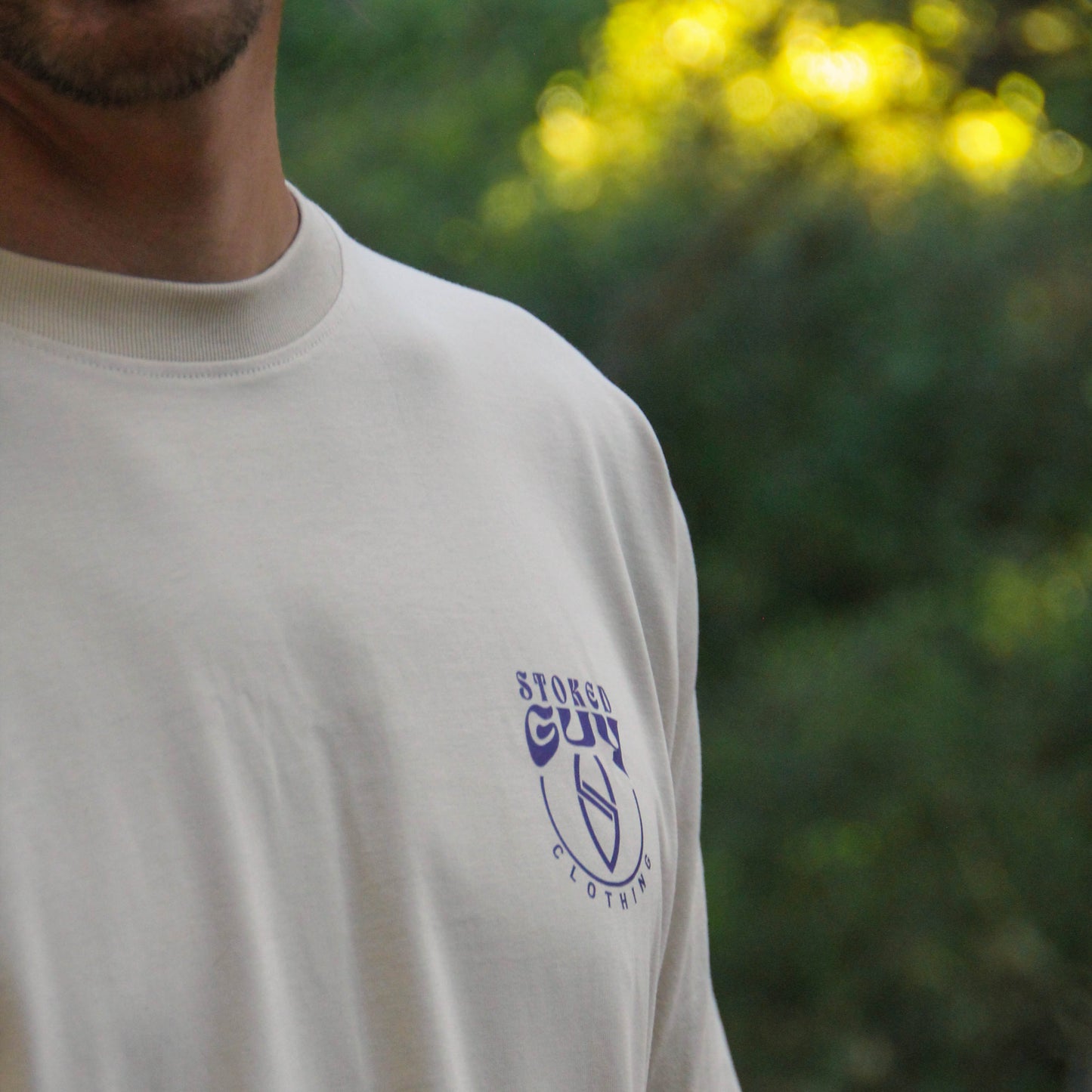 A close-up of a person wearing a cream-colored t-shirt with a blue 'STURGES CUP' logo on the left chest, set against a blurred green outdoor background.
