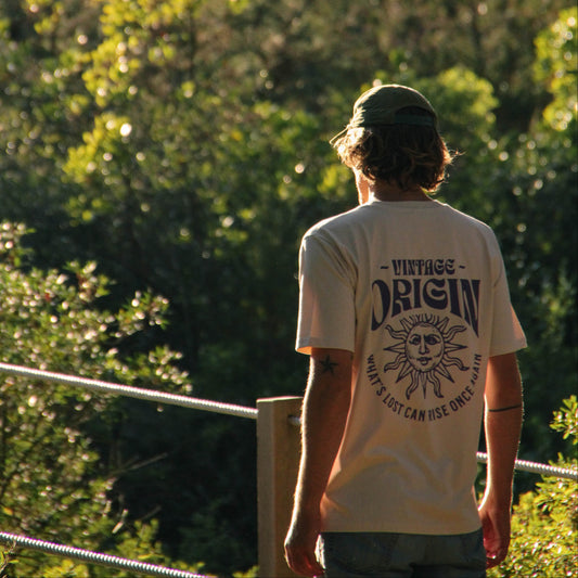 A man viewed from behind wearing a beige t-shirt with a dark graphic print that reads 'VINTAGE ORIGIN' and 'YOUR LIFE CAN RISE UP AS IT DEPARTS'. He is standing outdoors in front of a lush green forest background.