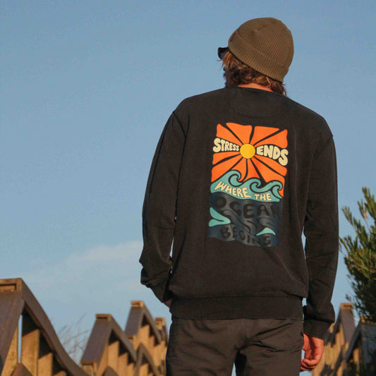 A man viewed from behind wearing a black crewneck sweatshirt with a large retro orange, green, and blue graphic on the back, paired with a brown beanie and dark pants, standing on a wooden staircase against a clear blue sky.